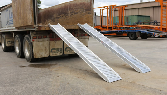 A pair of aluminium loading ramps attached to the back of a heavy-duty dump truck, set in an industrial yard with trailers and equipment in the background. The ramps provide easy access for loading and unloading heavy machinery.