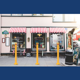 Four yellow removable bollards with red collars are installed in front of a charming bistro with striped red and white awnings, providing a protective barrier along the sidewalk