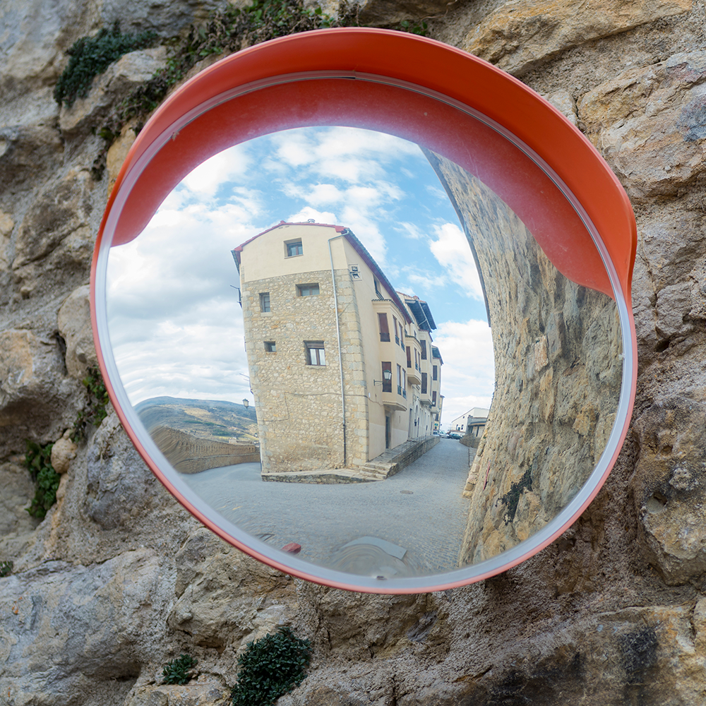 A convex mirror with a red border, mounted on a stone wall, reflecting a picturesque view of a narrow street and buildings. The mirror demonstrates its practical use in improving visibility in tight spaces.