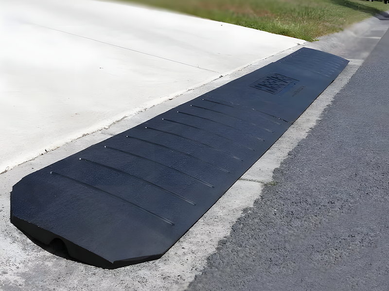 A long, heavy-duty black rubber kerb ramp placed at the edge of a driveway, providing a smooth transition from the road to the concrete surface. The ramp features a textured, non-slip surface with drainage channels and a slightly raised edge for added stability. It is designed for vehicles, wheelchairs, and trolleys to navigate kerbs safely. The surrounding area includes a neatly paved road, a bright concrete driveway, and a grassy verge in the background.