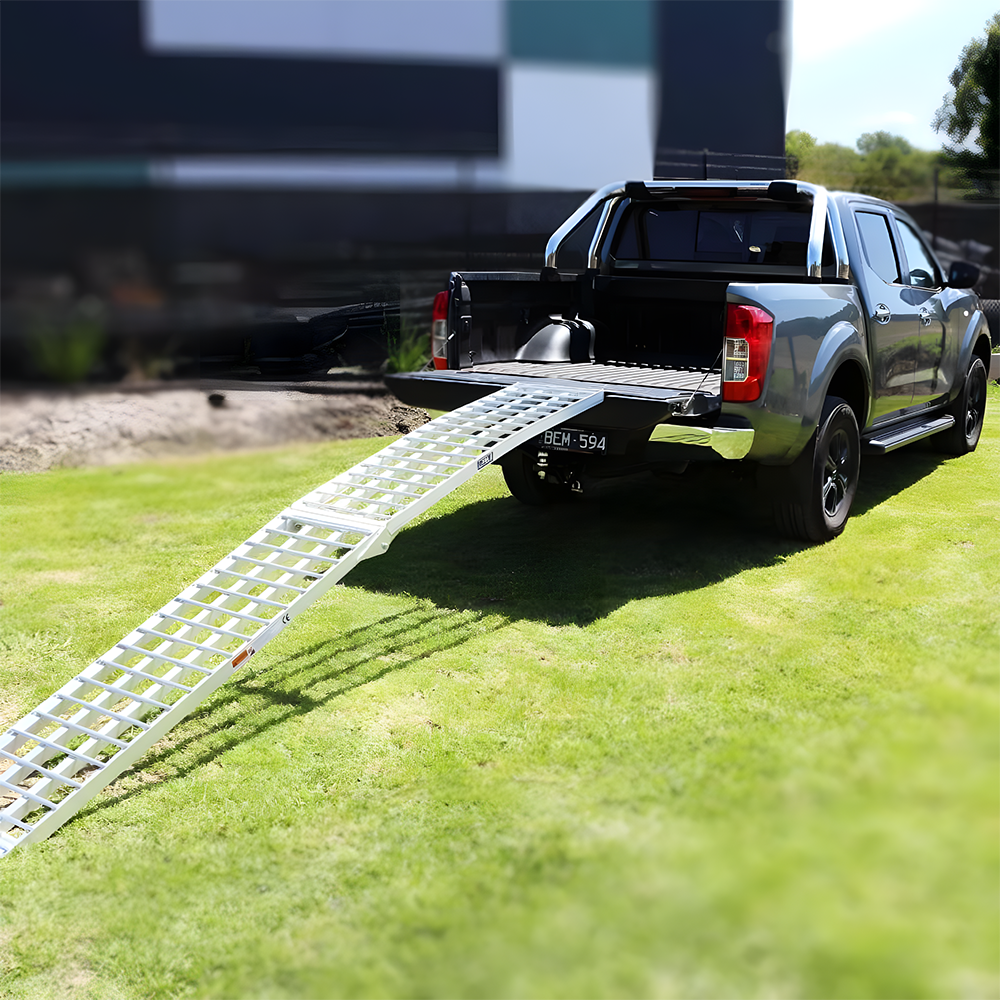 Reinforced aluminium loading ramp extended from the back of a vehicle, curving downwards to the ground on a sunny day with construction equipment in the background.