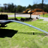 Reinforced aluminium loading ramp extended from the back of a vehicle, curving downwards to the ground on a sunny day with construction equipment in the background.