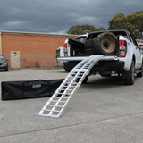 Standard aluminium loading ramp attached to the tailgate of a white pickup truck loaded with large tires, with a black Heeve carrying bag placed on the ground nearby