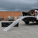 Standard aluminium loading ramp attached to the tailgate of a white pickup truck loaded with large tires, with a black Heeve carrying bag placed on the ground nearby