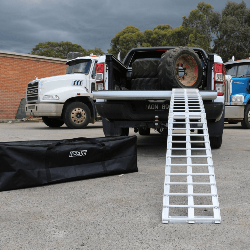 "A standard aluminium loading ramp extended from the tailgate of a pickup truck loaded with large tires, with a HEEVE branded carrying bag in the foreground."