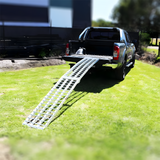 Reinforced aluminium loading ramp extended from the back of a vehicle, curving downwards to the ground on a sunny day with construction equipment in the background