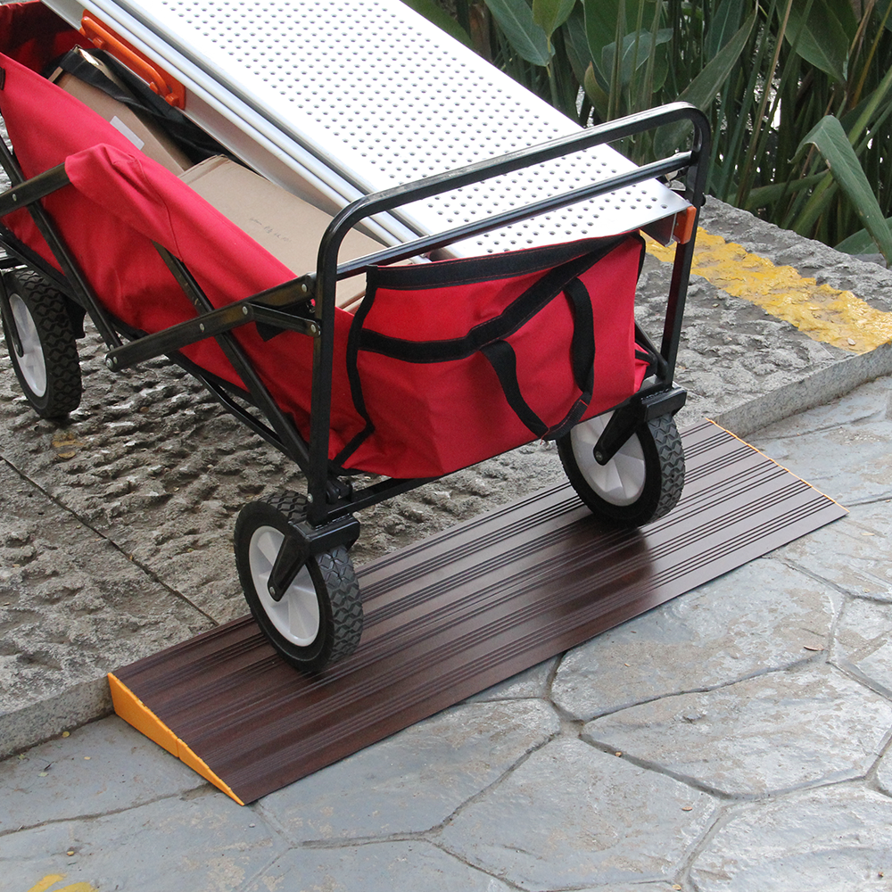 A red utility cart loaded with boxes and items, moving over a dark brown aluminium threshold ramp on an outdoor path.