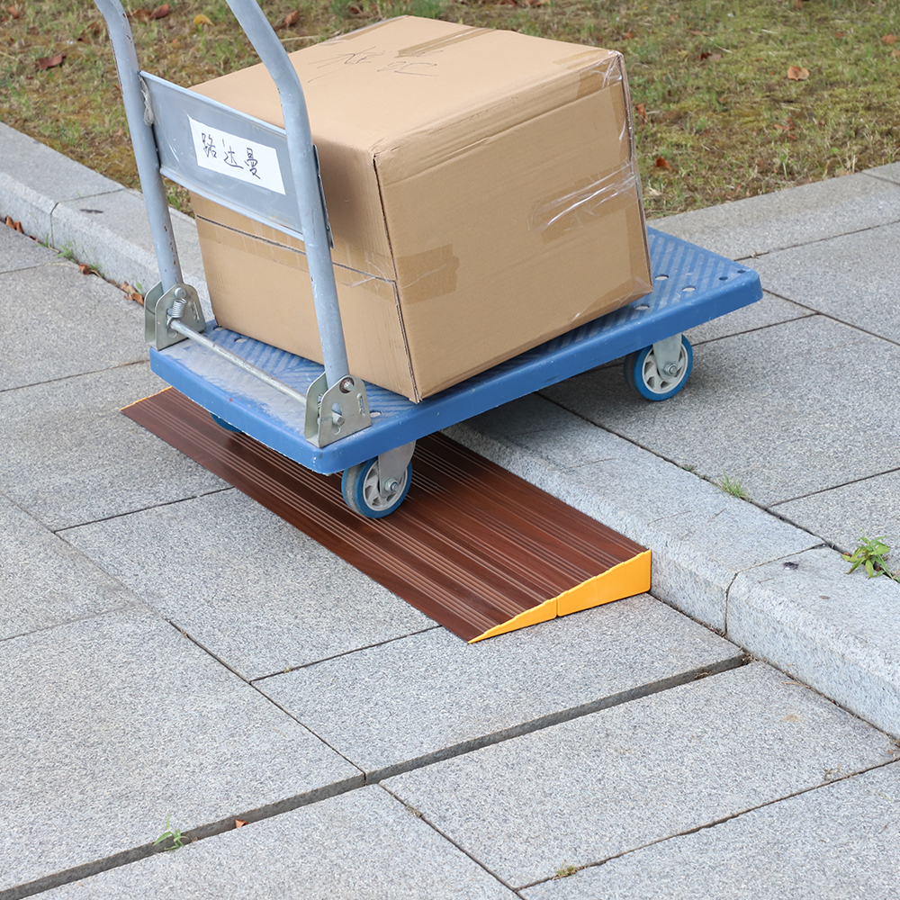 A blue trolley carrying a large cardboard box being wheeled over a brown aluminium threshold ramp positioned on a concrete pathway.