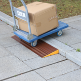 A blue trolley carrying a large cardboard box being wheeled over a brown aluminium threshold ramp positioned on a concrete pathway.