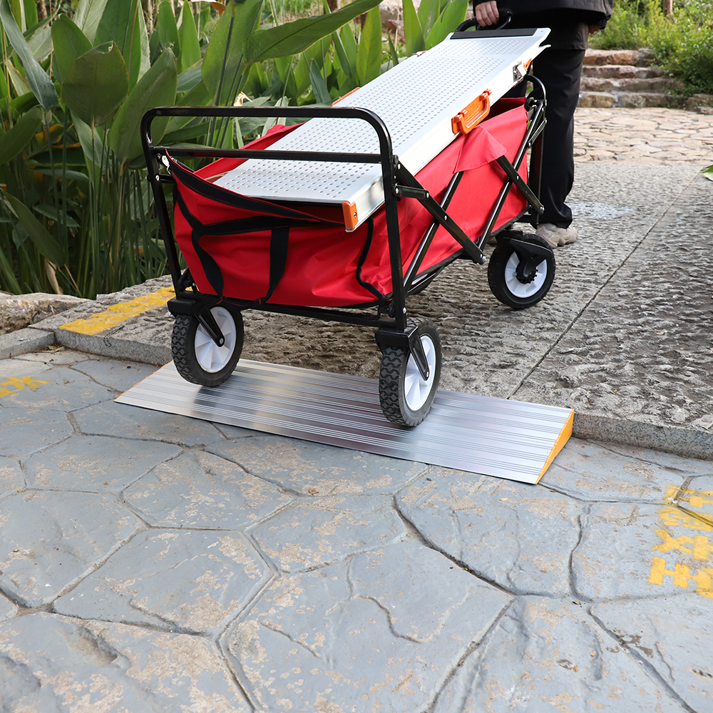 A person pushing a red utility cart over a silver aluminium threshold ramp on an outdoor pathway surrounded by plants.