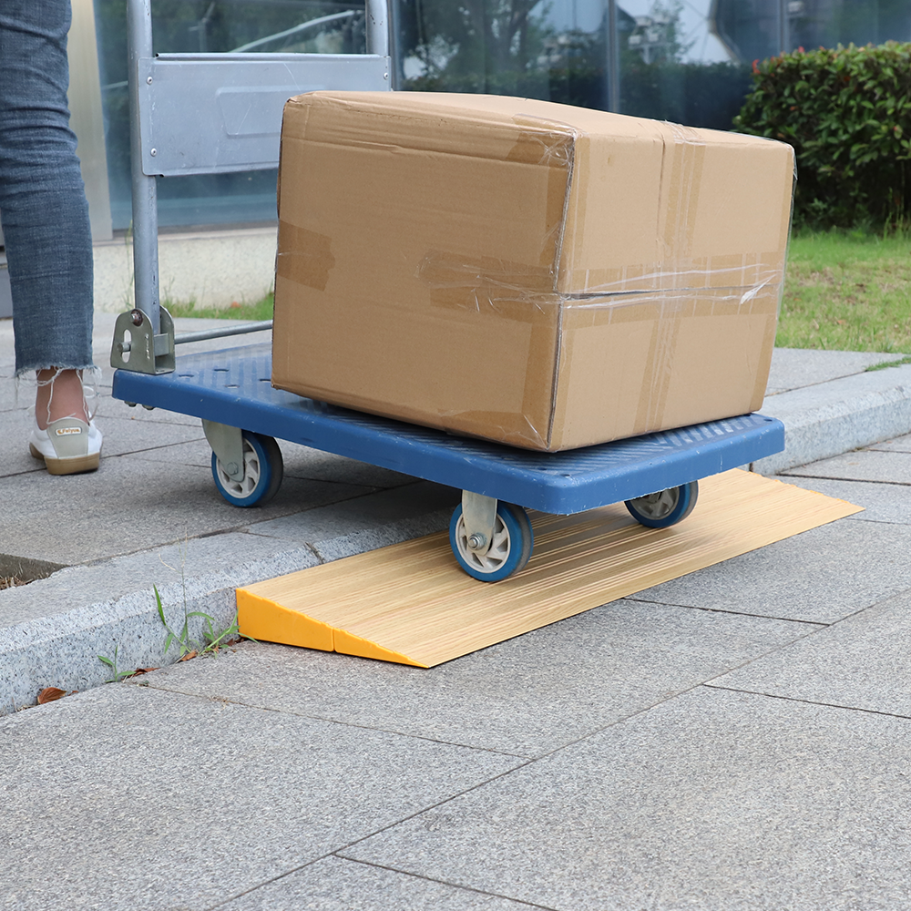 A blue trolley carrying a large cardboard box being wheeled over a wooden threshold ramp on a concrete pathway, with a person walking beside it.