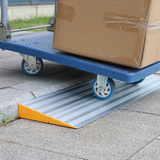 A heavy-duty aluminium threshold ramp placed on a kerb, with a blue trolley carrying a large cardboard box being wheeled over it