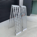 Two reinforced aluminium loading ramps folded and standing upright, showcasing their durable construction against a black garage door background