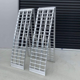 Two reinforced aluminium loading ramps folded and standing upright, showcasing their durable construction against a black garage door background