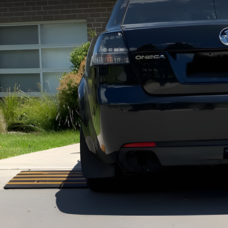 Rear view of a dark-coloured car parked on a driveway, crossing over a Heeve driveway kerb ramp with yellow stripes. The ramp is positioned at the edge of the driveway, facilitating a smoother transition over the kerb.