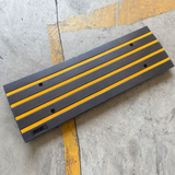 Close-up image of a Heeve driveway rubber kerb ramp with yellow stripes, designed for rolled-edge kerbs. The ramp is shown on a concrete floor, showcasing its textured surface and pre-drilled installation holes