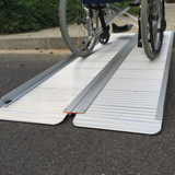 Side angle of a wheelchair rolling over the extended Heeve lightweight aluminium ramp on a road, showing its durability and sturdy build