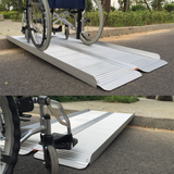 A wheelchair positioned on the Heeve folding aluminium ramp, providing easy access over a small kerb. Full view of the ramp extended on the pavement
