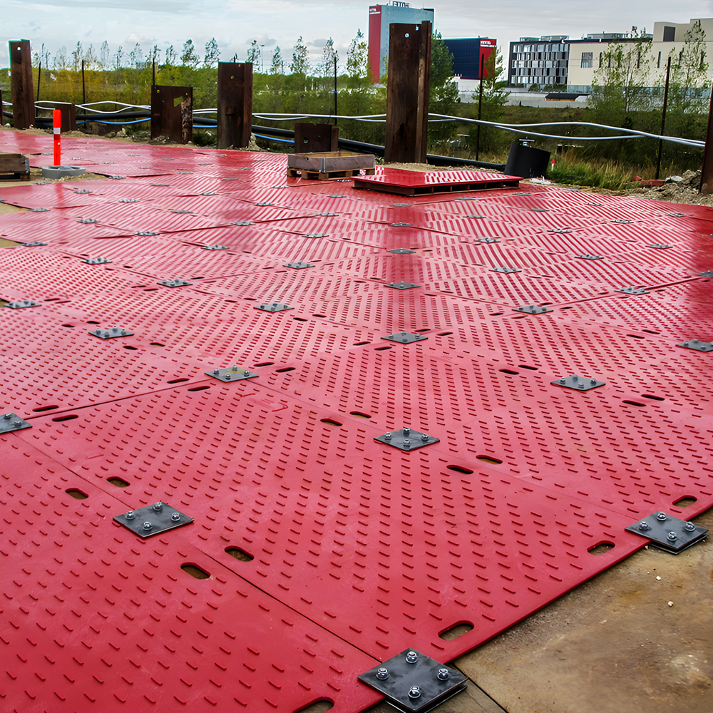 A construction site is shown with interlocking red ground mats, designed to create a stable surface, secured with metal connectors in a grid-like pattern