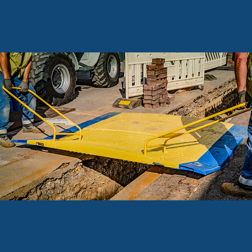 Two workers lifting a yellow LowPro road plate with handles to place it over a trench