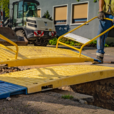 Workers using handles to lift a yellow LowPro road plate over a trench with construction vehicles in the background