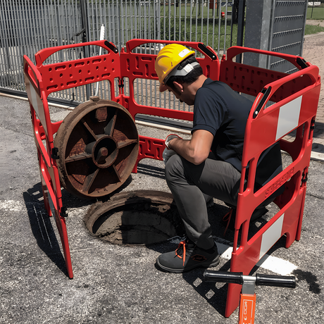 A construction worker in a hard hat kneeling down while inspecting an open manhole, surrounded by a red plastic four-sided barrier