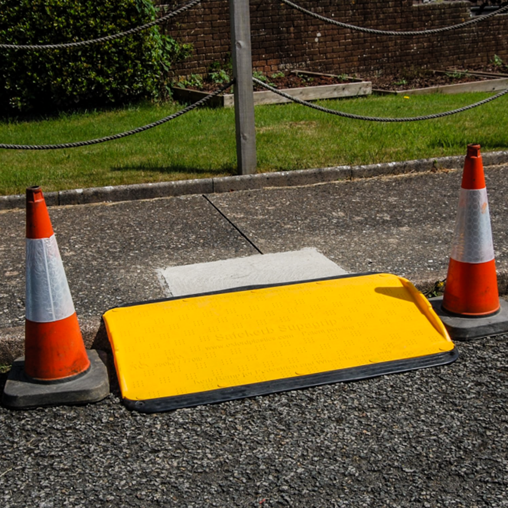 A yellow Supagrip Safekerb ramp positioned between two traffic cones on a sidewalk, ensuring pedestrian safety.