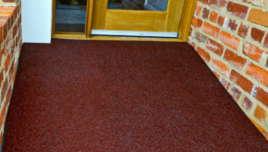 A durable rubber threshold ramp installed at the entrance of a brick home, providing a smooth transition over the doorway step. The ramp blends seamlessly with the rustic red brick walls and planter.